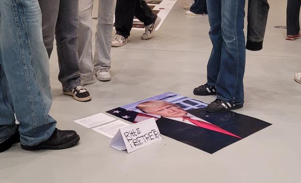 Auf dem hellen Boden einer Galerie liegt ein portraitfoto von Donald Trump.
Daneben ein Schild:" bitte betreten" und mehrere Beine in Jeans und Füße in Schuhen. Ein Fuß steht auf dem Foto.
