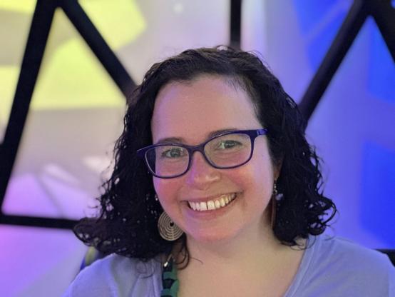 Head-and-shoulders color photo of writer Effie Seiberg, a fair-skinned woman with pink cheeks and curly black hair parted on one side and worn at shoulder length. She wears glasses with violet frames, large dangly earrings, and a light blue collarless knit shirt. She sits in what looks like an office chair with a black leather back in front of what might be a glass window in geometric shapes. She smiles broadly at the camera.