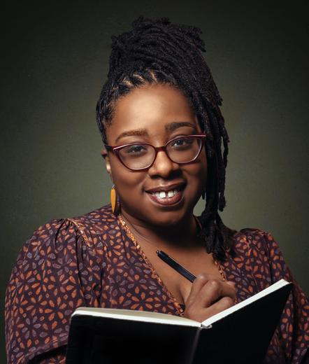 Head-and-shoulders color photo studio portrait of writer Erin Roberts, a Black woman with brown skin and black braids worn in a high ponytail that cascades to one side of her face. She wears cat-eye glasses with red frames, slender dangly earrings, and a brown-and-blue print dress or blouse in a simple repeating floral pattern. She stands before an olive-green background and holds a pen to an open notebook, looking directly into the camera and smiling warmly.