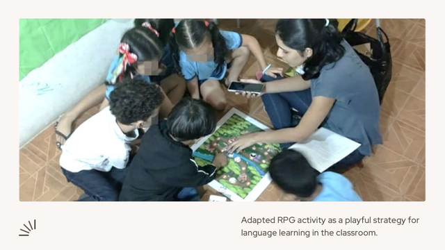A group of children sitting in a circle on the floor, engaged in an adapted RPG activity for language learning. They are focused on a colorful board game or map placed in the center. An adult facilitator is seated among them, holding a smartphone and guiding the activity. The setting appears to be a classroom with tiled flooring and chairs visible in the background. The text below the image reads: "Adapted RPG activity as a playful strategy for language learning in the classroom."