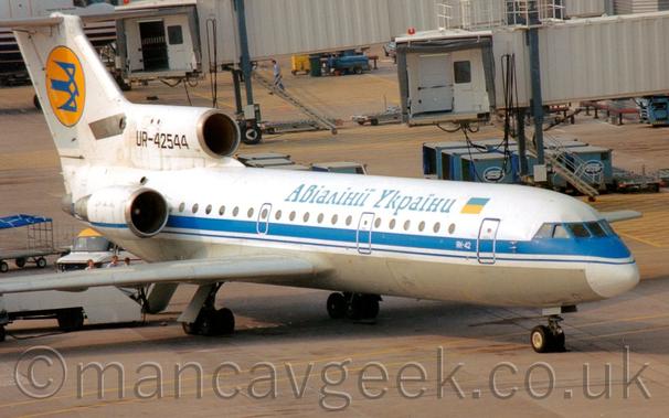 Side view of a white, 3 engined jet airliner with the engines mounted on the rear fuselage, parked facing to the right.
The plane has a grey belly, and a blue stripe running along the body under the cockpit windows.
There is blue text in a Cyrillic script on the upper fuselage, next to the flag of Ukraine (equal horizontal bars, blue on top, yellow below.)
There is a yellow circle on the tail, containing a stylised blue flying bird.
The registration "UR-42544" is on the centre engine intake, below and in front of the tail, in black.
Grey concrete apron fills the foreground, with white airbridges hanging out into space filling most of the background, assorted baggage carts, trucks, and other pieces of airport furniture scattered around the place.