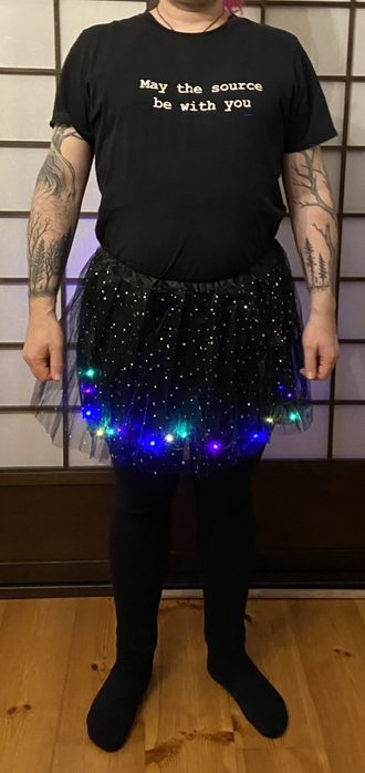 a person wearing a black t-shirt with "may the source be with you" print, black leggings or tights and a small black tutu skirt with glitter and LED lights