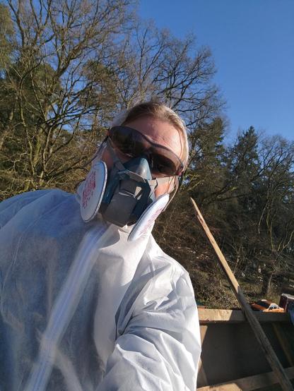 Selfie einer Person, die durch weißen Arbeitsschutzoverall, Atemschutzmaske und verdunkelte Schutzbrille kaum zu erkennen ist. Die Person steht auf einem Dach, im Hintergrund ist ein Waldrand und blauer Himmel zu erkennen