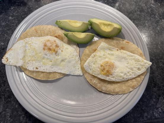 Gray plate on a black countertop, containing two roasted corn tortillas, side by side, with overeasy eggs on each, and 3 wedges of avocado just next to them (above on the shot)