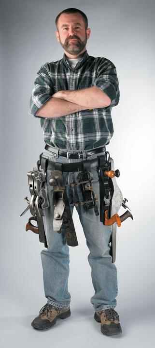 Shows a man in plaid shirt and worn jeans with the pouchless tool belt hanging off his waist. The tool belt holds many (a comically large number) hand planes, some of which extend to his knee.