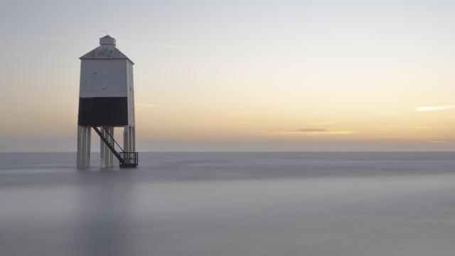 A wooden lighthouse on legs stands in shallow water just off the shore. The turbulent sea has been smoothed over with a 60 second exposure. The sun has set and the few clouds above the horizon are catching the last colour of the day.