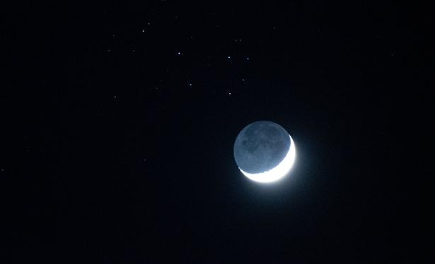 A crescent Moon against a black night sky with scattered stars in the background. The crescent is very bright white but the dark area of the surface can also be seen