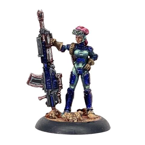 Soldier in futuristic body armor standing next to a giant rifle and holding it vertical as it rests on the ground on its butt. She has sparkly blue armor plates over a green-gray under armor. She has bright pink hair over goggles on her forehead. She stands on rocky red ground.