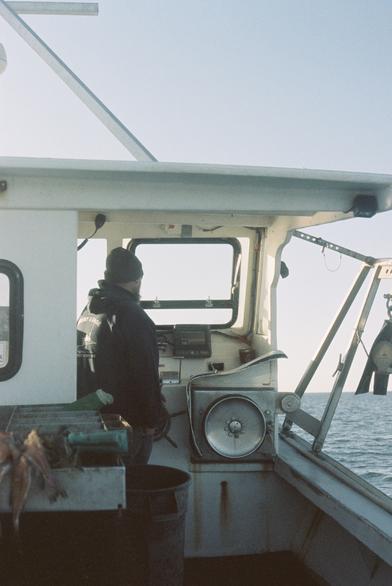 A 35mm film photo of a lobster boat taken standing at midship shooting forward toward the bow. Only a small portion of the boat is visible, including the rail, cabin top and windows, hauler, davit, and lobster tank. A man stands at the helm faced away from the camera.