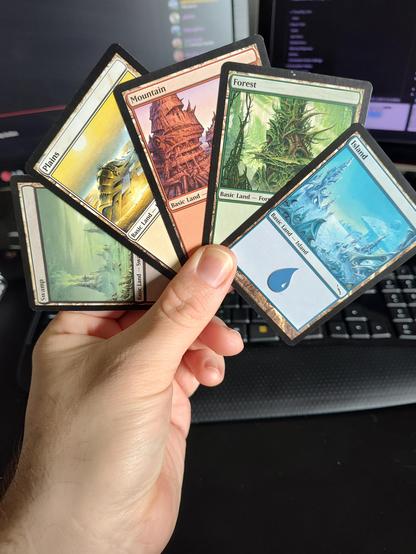 Photograph of my hand holding five Basic Land cards of the five Mana colors from the Magic the Gathering Mirrodin set.