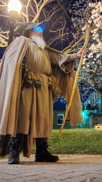 Middle aged man with long grey beard wearing Gandalf style outfit with hat and staff. Wearing funny light up techno glasses for April 1st. Blooming white tree in the background with street lamp.