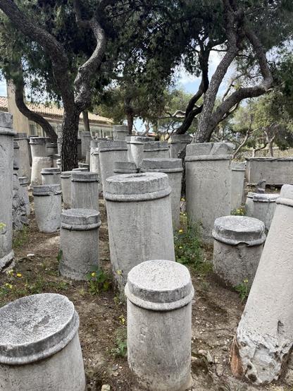 A cluster of columnar grave markers all grouped together at the archaeological site of the Kerameikos cemetery. They are about waist high and made of weathered marble, mostly unadorned, with a ring near the top and names carved into them.
Pine trees, and, in the background, a bit of the site’s lovely little museum.