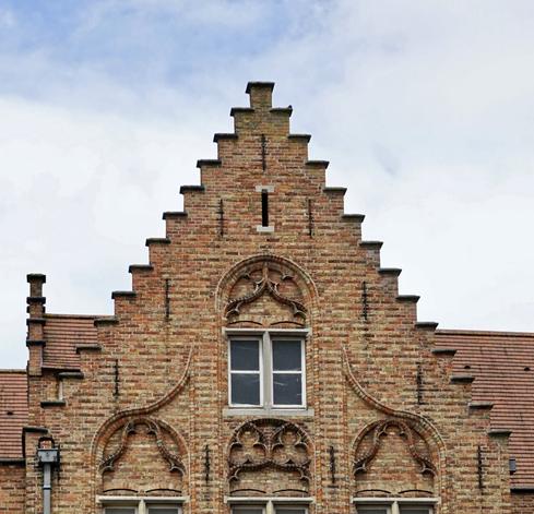 Photo du pignon crénelé en brique jaune d’un bâtiment ancien sur ciel bleu légèrement couvert. De jolis motifs en entrelacs décorent le dessus des fenêtres.
