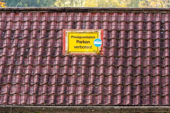 An einem Zaun, der direkt vor dem Dach eines tiefer gelegten Hauses steht, ist ein Schild auf dem "Privatgrundstück Parken verboten!" steht. Es wirkt, als bezöge sich das Schild auf das Dach.