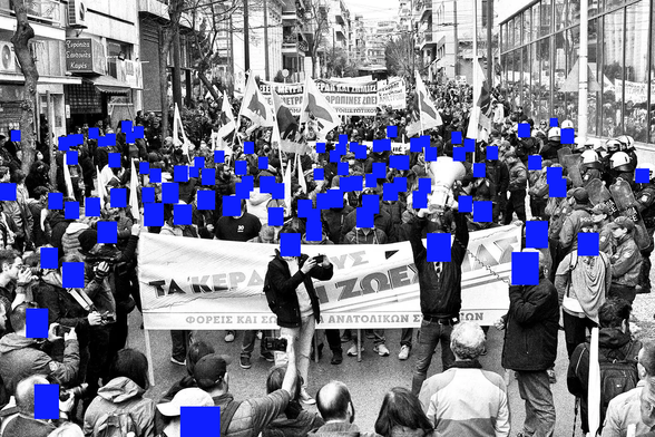 The image depicts a large crowd gathered in a street, likely during a protest or demonstration. The scene is in black and white, with blue squares obscuring the faces of many individuals. The crowd is holding banners and flags, with one prominent banner in the foreground reading "TA KEPARIZOGE" and "ΦΟΡΕΙΣ ΚΑΙ ΕΚΑΝΑΤΟΛΙΚΟΝ ΕΘΝΟΣ" (translated as "We Take Our Lives" and "Fronts and Anatolian Nation"). The crowd is diverse, with people of various ages and attire, some holding cameras and others raising their hands. In the background, there are buildings with signs in Greek, indicating the location might be in Greece. The atmosphere appears tense, with a line of riot police visible on the right side, equipped with helmets and shields. The overall composition suggests a significant public gathering with a strong sense of purpose.