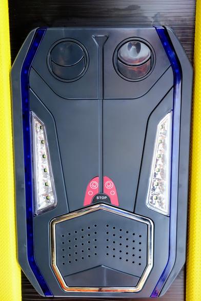 A close-up shot shows a rectangular, dark grey control panel, likely from the interior of a vehicle, possibly a bus given the "STOP" button. The panel has a symmetrical design. At the top are two circular air vents. Below these are two rectangular LED light strips, one on each side. In the centre is a red rectangular button with a white "STOP" inscription and two illuminated symbols above it. The bottom section features a speaker grille with numerous small perforations and a chrome-coloured border. The entire panel is outlined with a thin blue light strip. The background is dark, with a sliver of yellow fabric visible on the left and right edges.