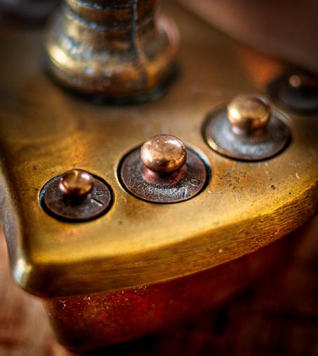 The photo shows a close-up of an antique apothecary scale with several small, round weights made of brass. The surface appears slightly worn and aged, with some areas showing oxidation and patina. The weights are arranged in a triangular pattern, and the metal surface has a warm gold tone with subtle reflections. The image features a shallow depth of field, blurring the background elements while highlighting the details of the metalwork. The combination of textures and tones gives the object a vintage or steampunk aesthetic.