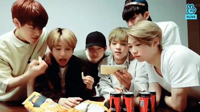Several skz members behind a table, with snacks and drinks in front of them. Seungmin looks mischievous as he snatches a snack away from Han's open mouth.