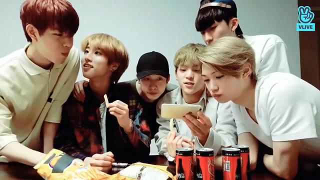 several skz members behind a table with snacks and drinks on it. Seungmin has just put a snack in his mouth and he has in hand in a bag, about to pull out another snack. Han gazes at Seungmin admiringly.