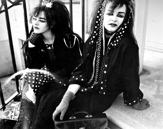 Black and white photo of Rose McDowall and Jill Bryson sat on stone steps. Jill mis nearer the camera, looking at us. Polka dot ribbons trail down from her hair. Rose looks into the distance, her chin resting on her fist. Both wear black coats and opera gloves, and have wing eyeliner and have probably used a whole eyebrow pencil between them