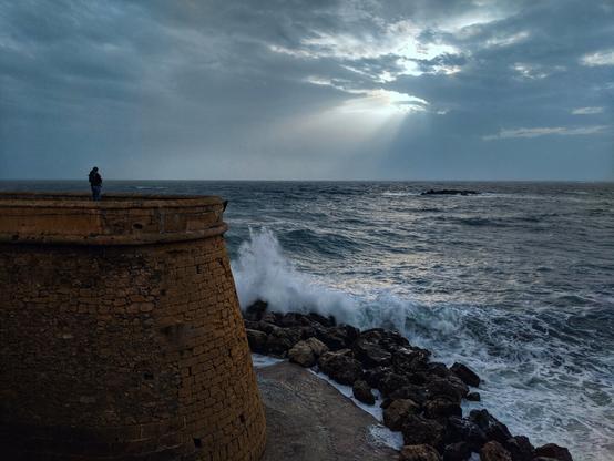 A wide shot captures a solitary figure standing atop a weathered, yellowish-brown stone bastion, overlooking a turbulent, dark grey sea under a dramatic sky. The sky is filled with heavy, dark clouds, but a break allows beams of sunlight to pierce through and illuminate a patch of the choppy water. White-capped waves crash against a rocky shoreline at the base of the bastion. The overall mood is one of brooding beauty and isolation.