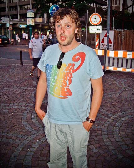 The image shows a man standing outdoors on a cobblestone street, with his hands casually placed in his pockets. He is wearing a light blue T-shirt with a colorful, rainbow-style text design that reads, "Tune in, drop out, stay high!" A pair of sunglasses hangs from the neck of his shirt. His expression is slightly surprised, with his mouth open.