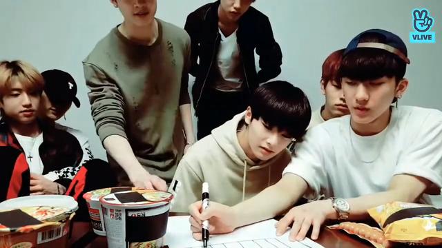 Skz members behind a low table. Changbin is drawing on paper. Lee Know sits to one side with Han on his lap. He's hugging Han from behind. Han has one hand raised to rest on top of Lee Know's. Han's expression is more settled and peaceful than his usual.