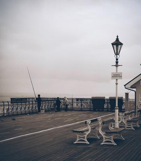 The image depicts a scene on a pier where three people are fishing. They stand near the edge of the pier, each with a fishing rod cast into the water. The pier is lined with white benches and a vintage-style street lamp with a sign that reads "FISHING PERMITTED".