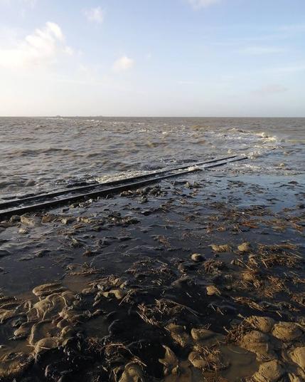 Eisenbahn-Gleise verschwinden in der anbrandenden Nordsee. Weit im Hintergrund sind Häuser auf den Warften der Hallig Nordstrandischmoor zu sehen.