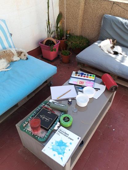 Foto de un rincón "salon" de una  terraza. Dos sofás de palet con cojines, una mesa baja. Encima de la mesa blocs de papel, una caja de pintura, pinceles, un añtavoz, un cenicero, ... En el fondo se ven varias plantas verdes en macetas.
