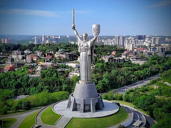 Das Bild zeigt mit der Mutter-Ukraine Statue in Kyjiw, auch bekannt als Mutter-Heimat, eine imposante Statue, die auf einem hohen Podest steht und ein großes Schwert in der rechten Hand und ein Schild mit dem Emblem des ukrainischen Tryzubs in der linken Hand hält. Die Statue ist mit silberfarbenem Edelstahl verkleidet und befindet sich inmitten eines grünen Parks. Im Hintergrund erstreckt sich die Stadt Kyjiw mit einer Mischung aus modernen Hochhäusern und älteren Gebäuden. Die Statue ist ein zentrales Element des Bildes und dominiert die Szene. Der Himmel ist klar und blau, was die Szenerie noch beeindruckender macht. Die Statue ist ein Symbol für Stärke und Frieden und ist ein bekanntes Wahrzeichen der Stadt.