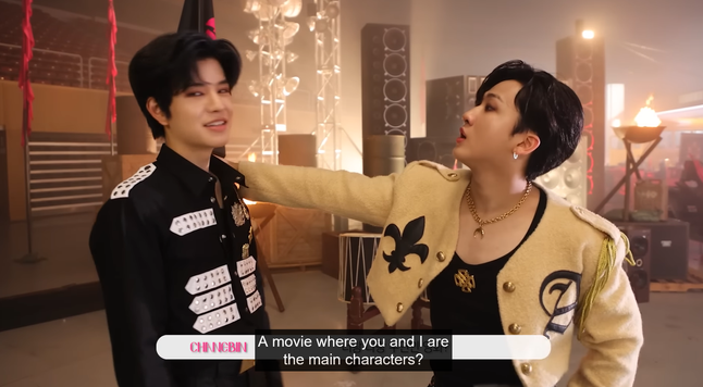 Seungmin and Changbin on the set of LALALALA MV. Seungmin's looking at the camera while Changbin poses like a sexy romantic lead, with his head tilted back and his hand on Seungmin's shoulder. Caption: "A movie where you and I are the main characters?"