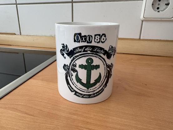Foto einer weißen Tasse. Oben Aufschrift „Oxo 86“, grüner Anker, Aufschrift „auf die Liebe und auf die Sehnsucht“