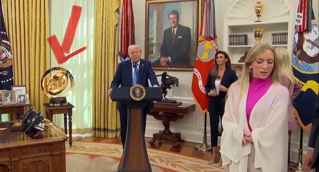 Donald Trump und ein paar Angestellte im Oval Office, im Hintergrund auf einem extra-Tischchen eine große sphärenförmige Skulptur ganz in Gold, markiert durch einen handgemalten roten Pfeil.
