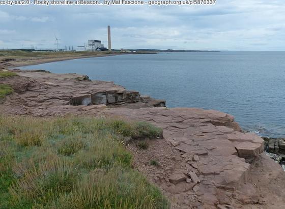 Low rocky shoreline on the left, and the sea on the right. In the distance a power station. Chimney