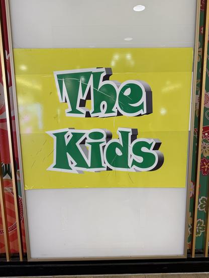 A brightly colored sign featuring the words "The Kids" in green and black text against a yellow background. The sign appears slightly damaged, with creases and scratches. It is displayed on a wall with a decorative, patterned background.