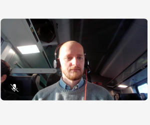 Das Bild zeigt mich mit kurzen Haaren und Bart, wie ich in einem Zug sitze. Ich trage Kopfhörer mit einem Kabel. Im Hintergrund sind die Innenräume des Zuges zu sehen, einschließlich der Sitze, der Fenster und der Deckenleuchten. Ein weiterer Passagier ist teilweise sichtbar. Die Atmosphäre wirkt ruhig und entspannt.