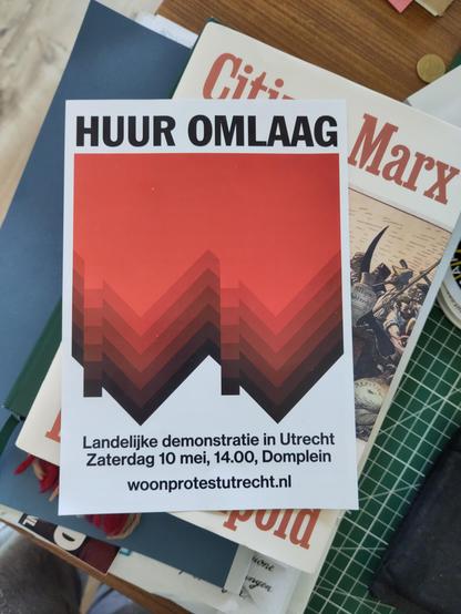 een flyer ligt bovenop een boek over Marx. op de flyers in koeien letters: "HUUR OMLAAG" en daaronder "Landelijke demonstratie in Utrecht, Zaterdag 10 mei 14.00 Domplein, woonprotestutrecht.nl"