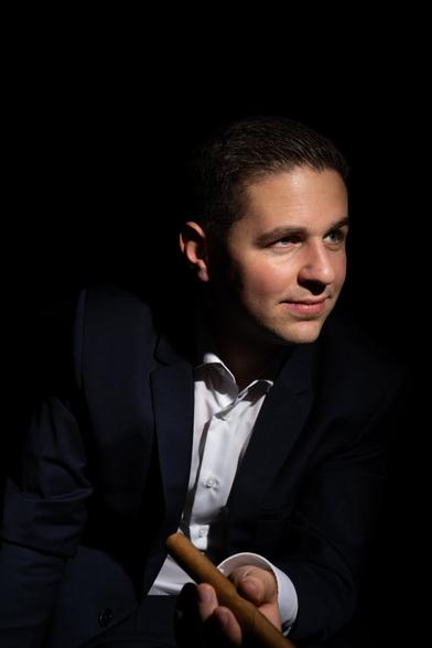 A person in a dark suit jacket and white shirt is seated against a black background, holding a cigar in their left hand and appearing slightly angled to the right. One side of the face is illuminated, while the other is obscured by shadow.