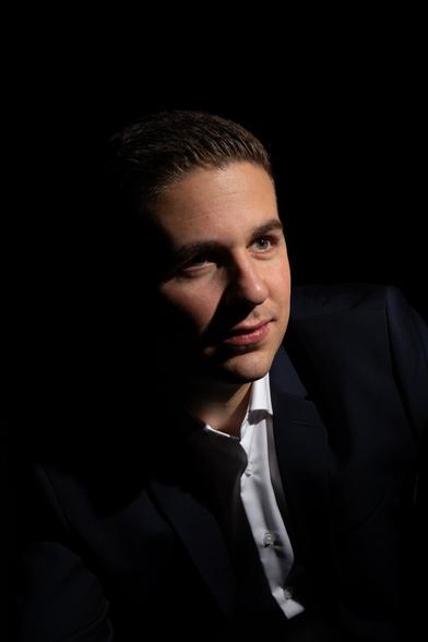 Portrait photo of a person in a dark suit jacket and white shirt, shown against a completely black background. The lighting illuminates the right side of the face, leaving the left side mostly in shadow. The individual is looking slightly to their left, with a thoughtful expression. The overall mood is dramatic and low-key, emphasizing contrasts between light and darkness.