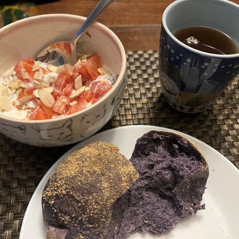 A small white plate holds a purple roll that is torn in half. Above the plate is a bowl half filled with a creamy speckled portage topped with red orange pieces and almond slices. A blue cup decorated with a winter theme has a black tea