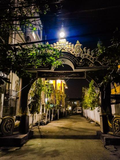 Photo shows an alleyway by night, with a gate with classical figures of the type typical for Burmese history and buddhist traditions. Some lights shows part of the smal way, and some of the old facades. It is a peaceful evening in an old city.
