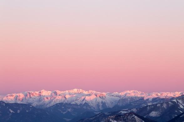 This image shows a mountain range with snow-covered peaks bathed in pink light, likely from a sunrise or sunset. The sky above is a gradient of soft pink hues, creating a serene and picturesque scene.