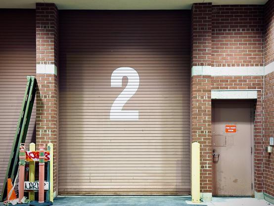 A large, reddish-brown garage door with a white "2" painted in the middle stands tall under a white ceiling at night, flanked by ladders/cones/caution signs on the left and a "FIRE RISER ROOM" door on the right.