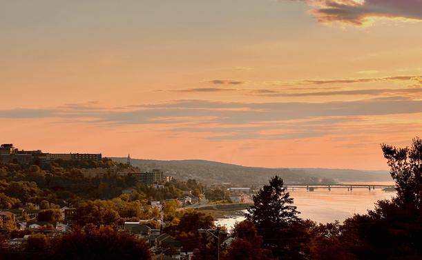 A scenic view of the Saguenay River at sunset, with soft orange and pink hues in the sky. In the foreground are trees and buildings on a hillside, while the river reflects the colours of the sky. A bridge crosses the river in the distance, and the landscape is dotted with greenery and houses.