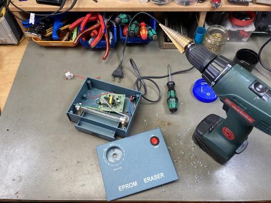 My cheap EPROM eraser on my workbench with the lid removed. A new power switch was added and the mechanical timer was removed. A cordless drill with a step drill bit is sitting next to it on the desk.