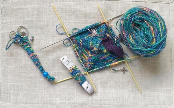 Une paire de ciseaux de broderie dans son étui, un mètre souple blanc dans son tube et le le début d'un béret tricoté à cinq aiguilles sur un fond écru. Le fil utilisé pour tout est multicolore avec une dominante verte.
Sur le fond du béret on voit des marqueurs de tricot dont un est une chaussette miniature violette.