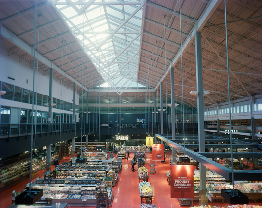 A view from the mezzanine overlooking the fresh produce department of a very large grocery store, the arched ceiling skylight provides much of the light and the light and sign fixtures hang from long vertical poles. Shoppers stroll the blood orange tiled floor.