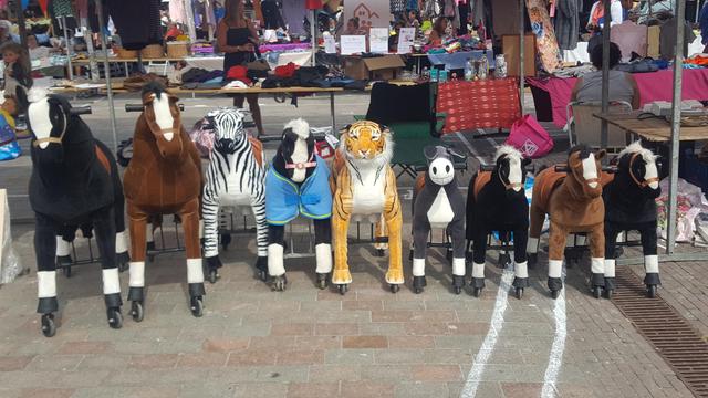 A line of walking bike style animals on large casters in front of a flea market stall. From left to right: a big black and a big brown horse, a medium sized zebra, black horse in blue blanket and tiger and small donkey and three small horses: a brown flanked by a black one on both sides.