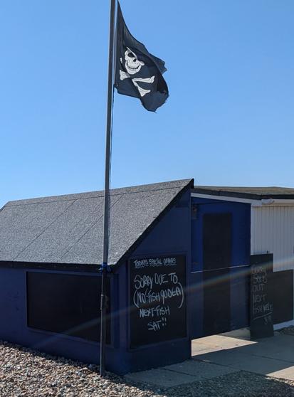 A fisherman's guy on a shingle beach with a skull and crossbones flag flying above it. A chalk board on the hut reads 
"SORRY DUE TO NO FISH QUOTA)

NEXT-FISH

SAT!!"